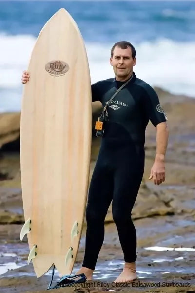Surfer standing beside a Riley balsa Fish surfboard on the beach