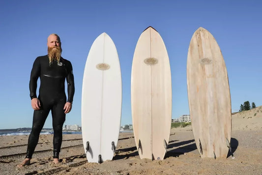 Mark Riley standing beside a line-up of handcrafted balsa surfboards on the beach